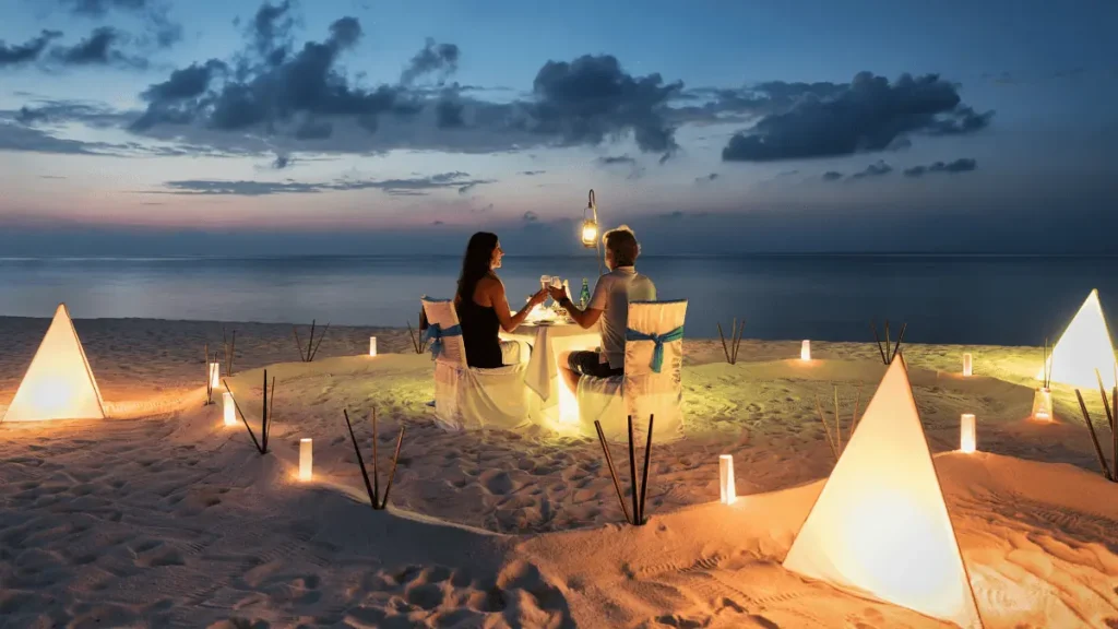Pareja cenando en la playa al atardecer con iluminación romántica y decoración con velas.