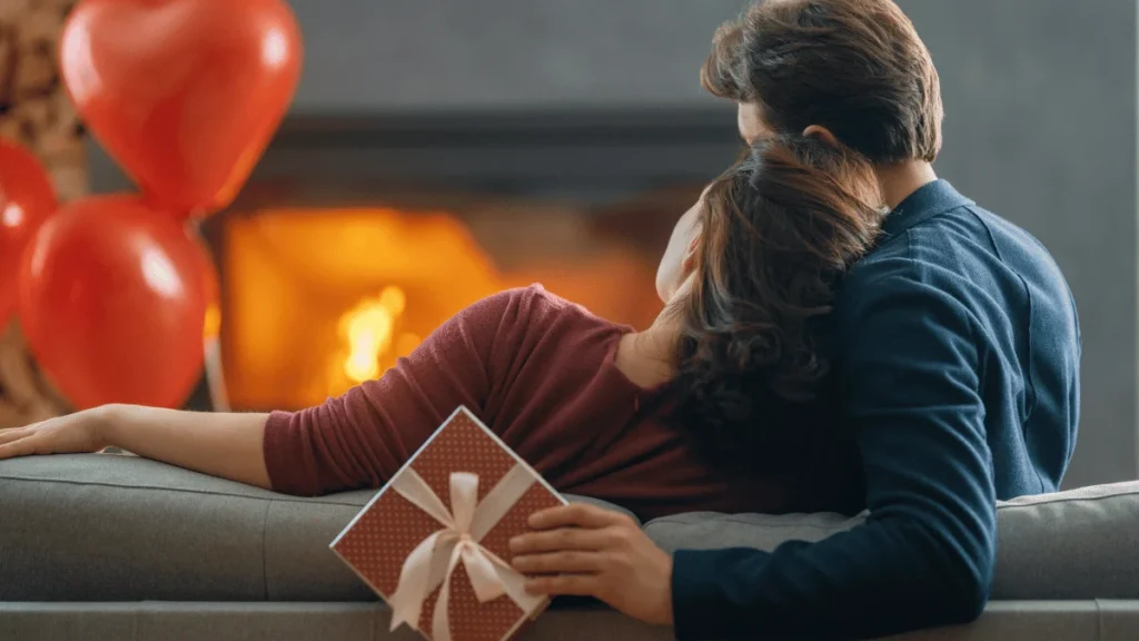 Pareja sentada en un sofá frente a una chimenea, con globos rojos en el fondo. La mujer sostiene un regalo envuelto con un lazo blanco mientras se recuesta sobre el hombro del hombre, creando un ambiente íntimo y acogedor.