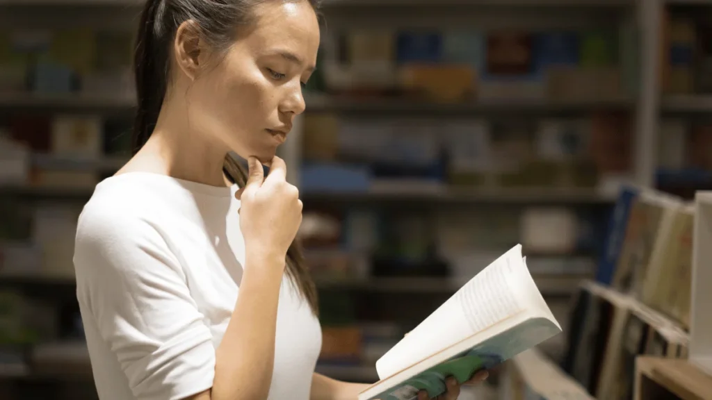 Mujer joven pensativa leyendo un libro en una biblioteca, rodeada de estanterías llenas de libros. Ambiente de estudio y reflexión.