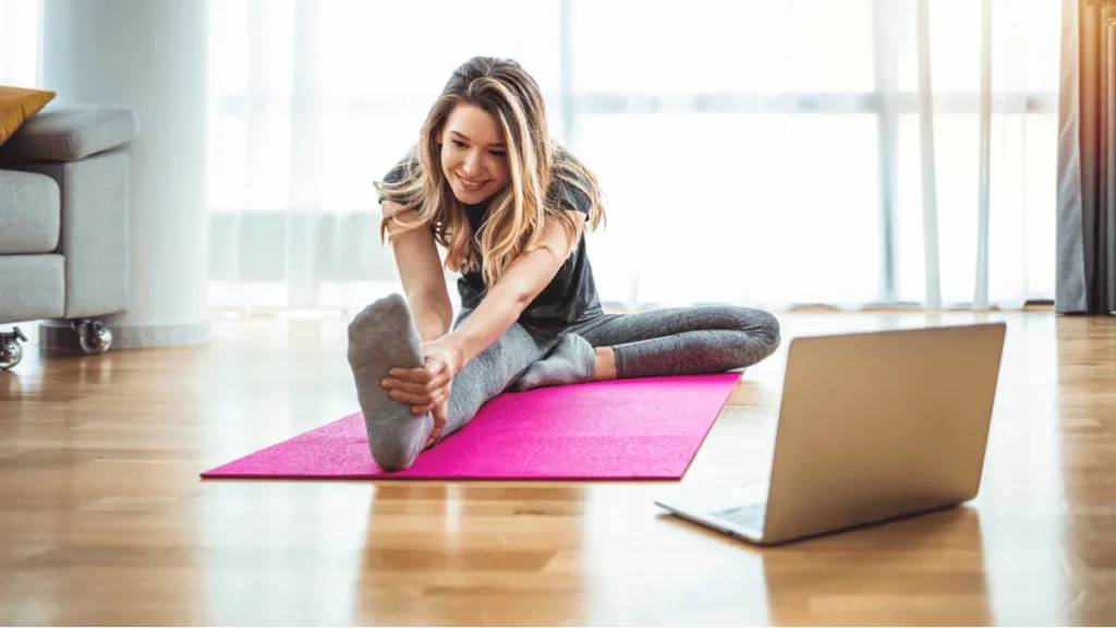 Mujer joven sonriendo mientras hace estiramientos en una esterilla de yoga rosa en casa, siguiendo una clase en línea en su laptop. Ambiente de ejercicio y bienestar en el hogar.