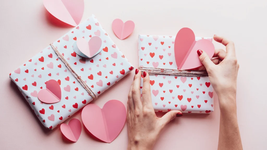 Manos envolviendo regalos con papel de corazones y corazones de papel.