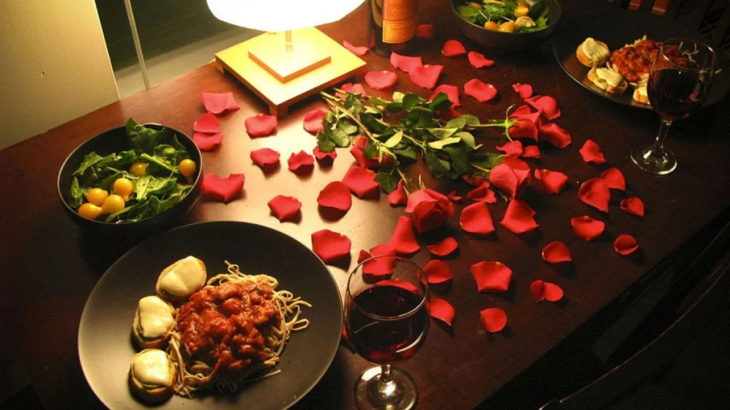 Cena romántica con pétalos de rosa, espagueti, vino y ensalada sobre una mesa de madera.