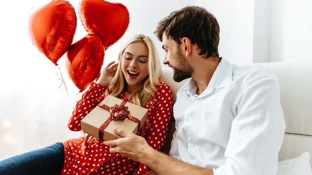 Pareja feliz intercambiando un regalo con globos rojos en forma de corazón en el fondo.