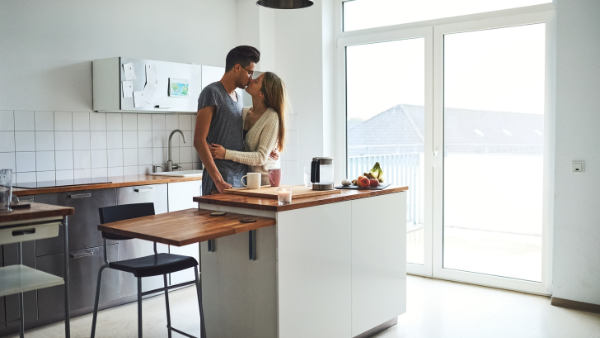Una pareja joven se besa en una cocina moderna y luminosa. La mujer abraza al hombre mientras se inclinan sobre una isla de cocina de madera, disfrutando de un momento íntimo juntos.