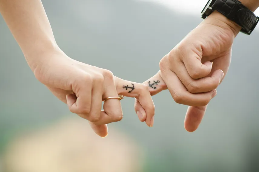 Manos de una pareja entrelazadas mostrando tatuajes de anclas en los dedos, simbolizando un vínculo fuerte y duradero.