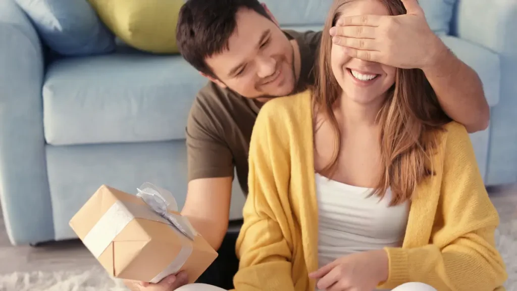 Hombre tapándole los ojos a una mujer y sosteniendo un regalo envuelto con una cinta blanca, ambos sonrientes y sentados en una sala acogedora.