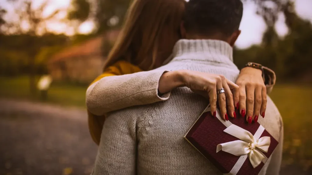 Pareja abrazándose en un entorno otoñal, con la mujer sosteniendo un regalo envuelto con una cinta blanca detrás del hombre.